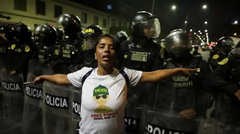 REUTERS/Sebastian Castaneda A protester, wearing a white T-shirt with the slogan "police, you're poor, same as me", spreads her arms out at her side as she stands in front of a line of riot police holding shields. in Lima, Peru, on 28 September, 2025.