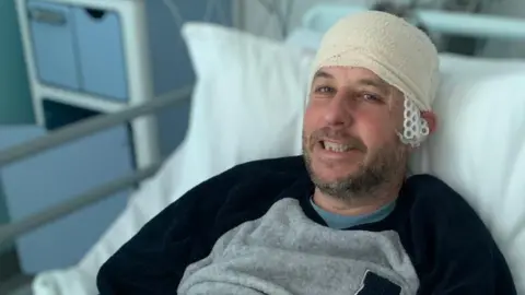 Cancer Research UK handout Alex Davies lying in a hospital bed. He has brown stubble with a bandage around his head. He is wearing a black and grey top. Alex is smiling.