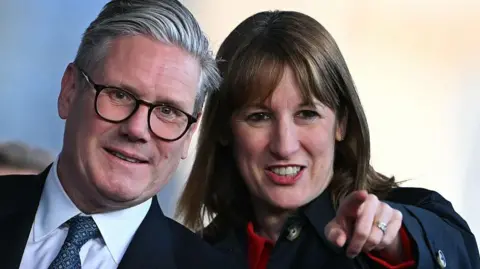 WPA Pool/Getty Images Prime Minister Sir Keir Starmer wearing glasses and a suit, standing next to Chancellor Rachel Reeves who is wearing a dark coat over a red top. She is pointing into the distance and they are both looking in that direction and smiling