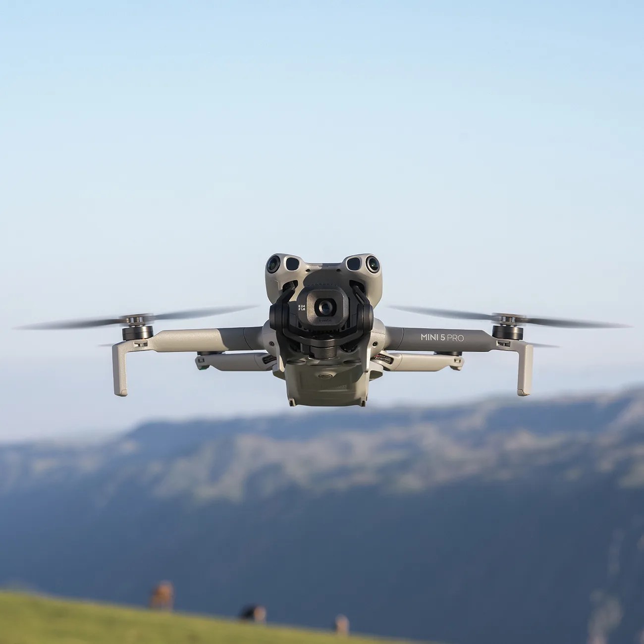 A DJI Mini 5 Pro drone in flight, shown from the front with its camera and gimbal visible. The drone's propellers are spinning, and a blurred mountainous landscape is in the background under a clear sky.