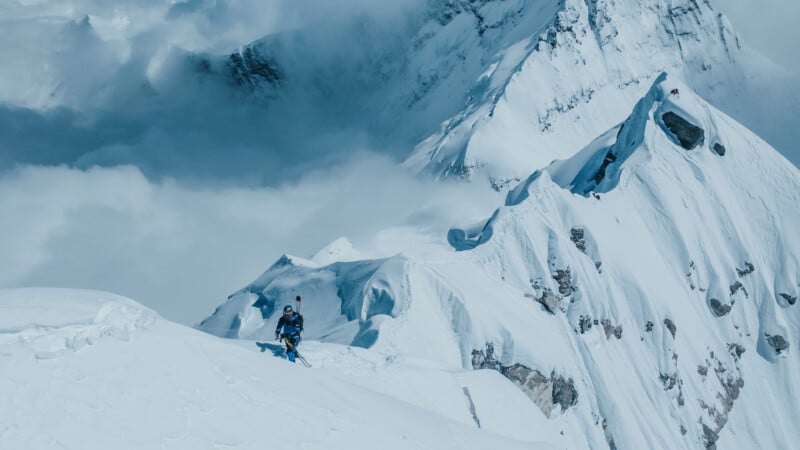 A lone mountaineer in winter gear climbs a snowy, steep ridge surrounded by dramatic, snow-covered peaks and swirling clouds high in the mountains.
