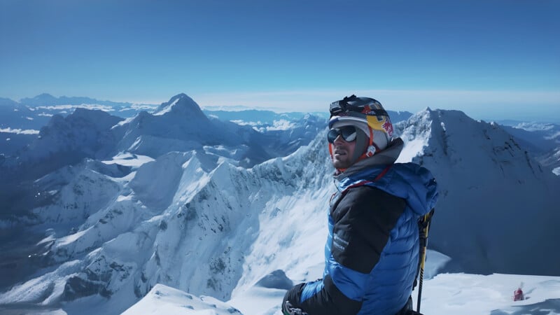 A climber in a blue jacket, helmet, and sunglasses sits on a snowy mountain peak, gazing at the surrounding snow-covered mountain range under a clear blue sky.