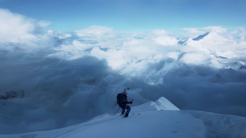 A lone climber in winter gear stands on a snowy mountain ridge above the clouds, surrounded by dramatic peaks and a vast, cloudy sky.