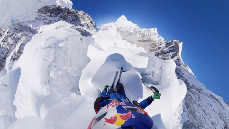 A climber wearing a Red Bull helmet ascends a snow-covered, steep mountain ridge, using climbing poles. Jagged peaks and a clear blue sky are visible in the background. The image is shot from the climber’s perspective.