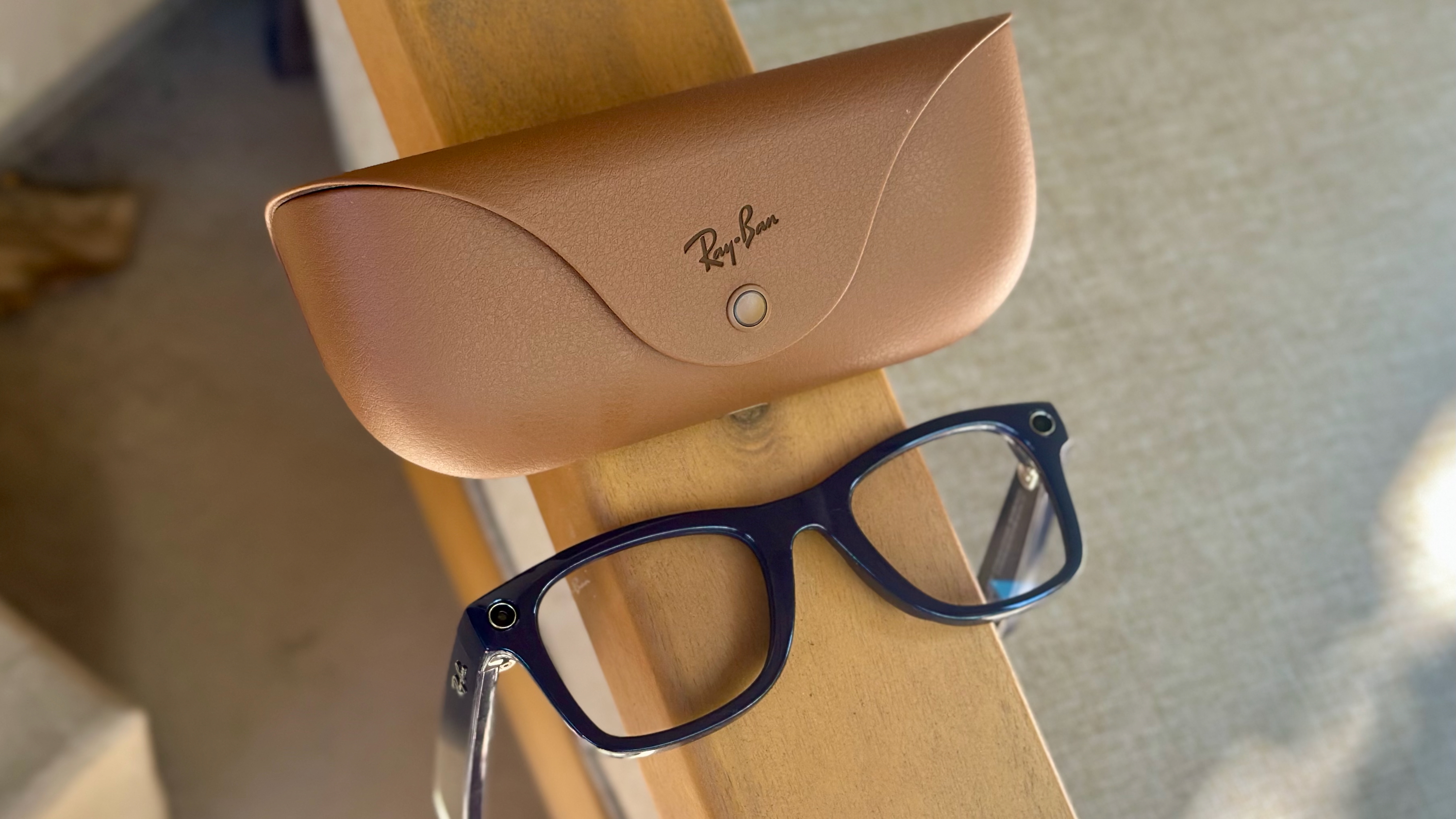 Close-up, top-down photo of the Shiny Cosmic Blue Ray-Ban Meta (Gen 2) Wayfarer glasses laying lenses up, arms down on an armrest, with the official brown Ray-Ban charging case behind it.