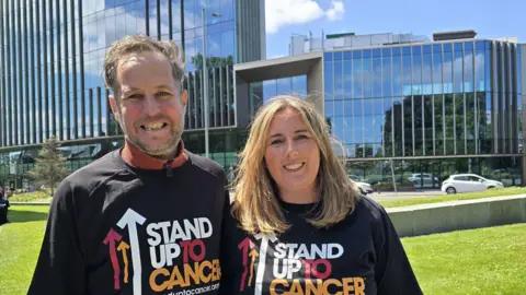 Cancer Research UK handout Alex, who has brown hair and beard and Emma Davies, with long blond hair, standing outside the entrance to Manchester Cancer Research Centre wearing t-shirts which read Stand Up To Cancer. They are both smiling. It is a sunny day.