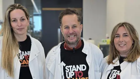 Cancer Research UK handout Left to right: Prof Petra Hamerlik, Alex and Emma Davies in a lab in the Manchester Cancer Research Centre. They are all wearing white coats and smiling. 