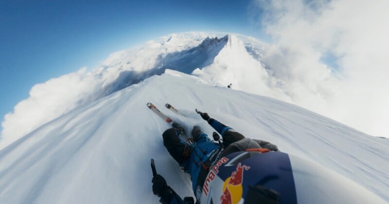 A skier in winter gear, seen from a first-person view, speeds down a narrow, snowy mountain ridge surrounded by clouds and distant peaks, creating a dramatic sense of height and adventure.